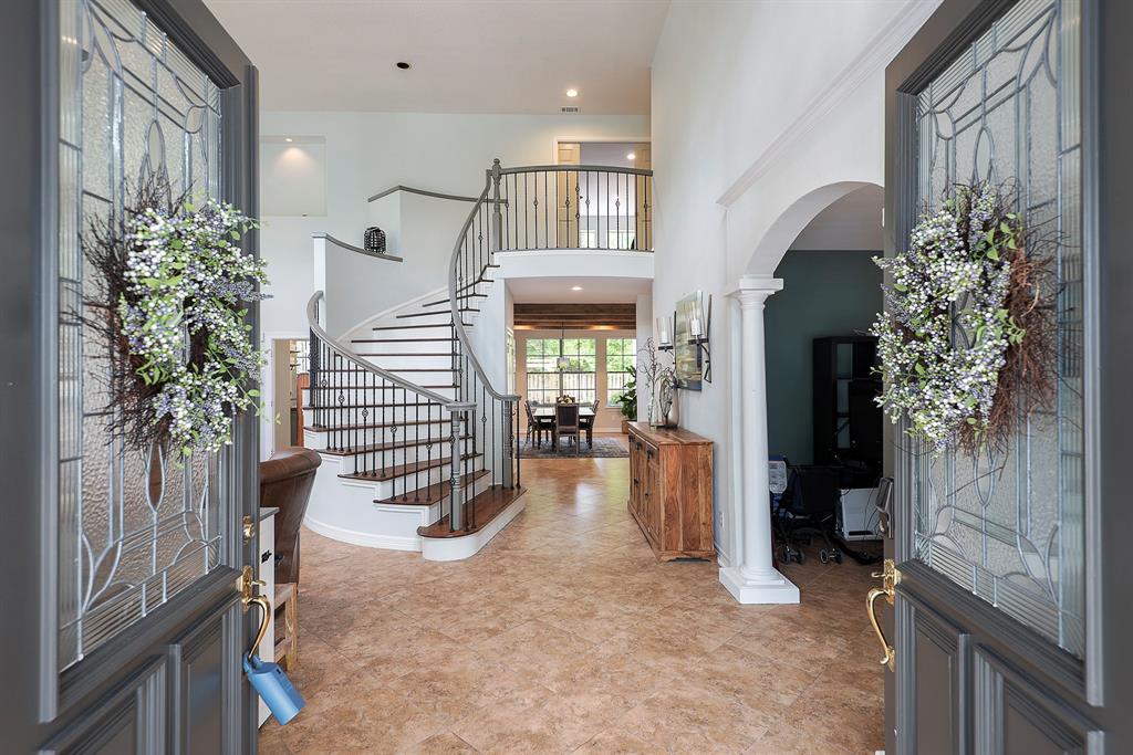 Open double doors framed by decorative glass panels, each with a spring wreath, leading into a bright foyer with a curved staircase and tiled floor.